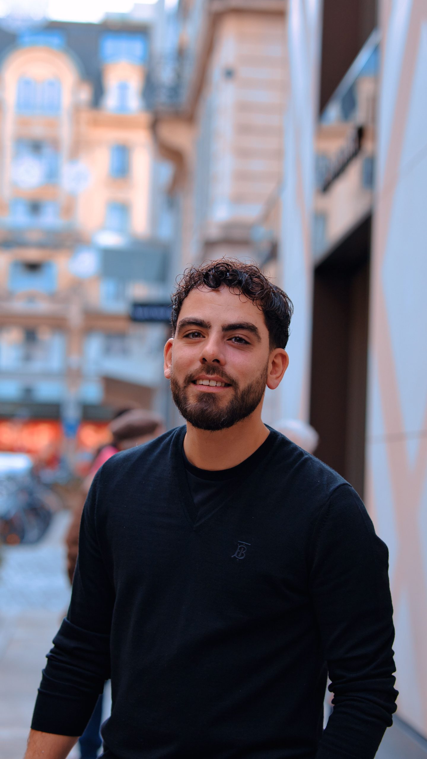 Smiling man in a black sweater posing outdoors with buildings in the background.